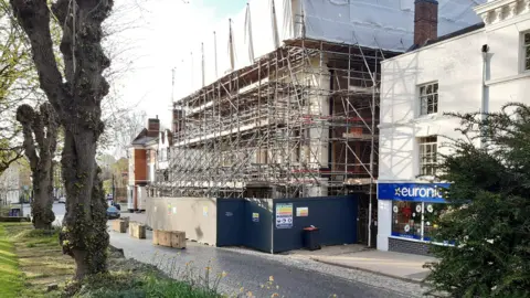A hotel building covered in scaffolding and with a large barricade set up at the base. It is pictured between other buildings at the side of a street.