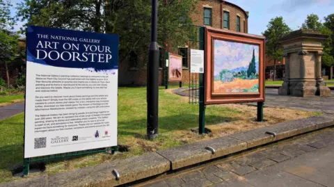 An interpretation board next to a replica artwork. The board has a dark blue top with white writing that reads The National Gallery Art on Your Doorstep. The artwork and board have been installed on a patch of lawn 