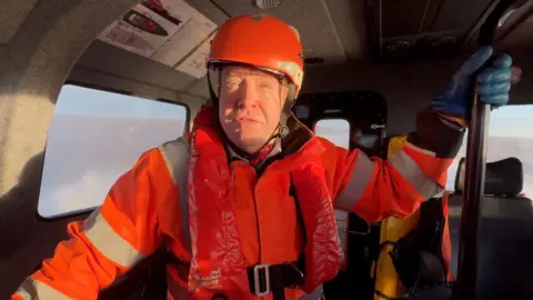 A man wearing an orange helmet, protective gear, blue gloves and a red life jacket stands inside one of the new pilot launches with the North Sea behind him.