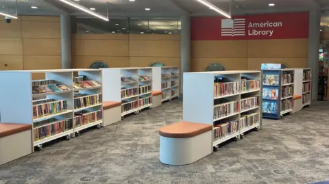 Owen Sennitt/BBC Rows of book shelves, and colourful books, which are about the chest height of an adult. There is a grey patterned carpet. There are wood panels on a wall.