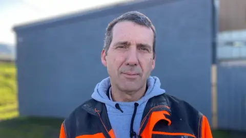 A man wearing a grey hoodie with a black and orange coat on. He is looking directly into the camera and has short hair. He is standing outside on grass with a grey wall behind him. 