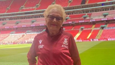 Bantam of the Opera An older lady with short, light brown hair stands in front of the pitch of a football stadium.