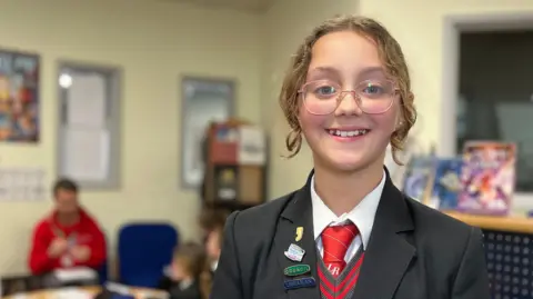 Beatrice is in a dark school uniform, including a black blazer, white shirt and red tie. She is standing smiling in a classroom, with a small group working at a table behind her, and bookshelves in the background.