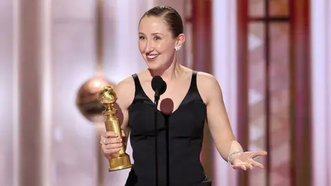 Getty Images Doherty standing on a stage at the awards ceremony. She is holding a Golden Globe trophy in the left hand while speaking into a microphone positioned on a stand in front of her. She is wearing a sleeveless black dress with a fitted bodice and a decorative bow detail near the waist. A bracelet is visible on the right wrist, and earrings can also be seen. The background features a patterned wall with vertical panels and several spherical decorative elements.