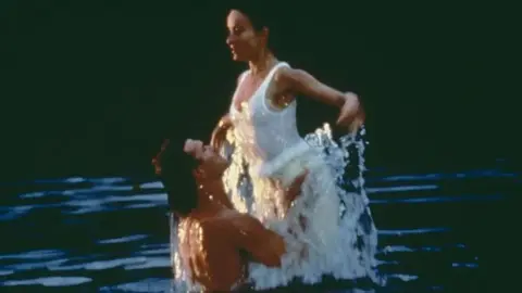 Getty Images A still shot from the film Dirty Dancing. Patrick Swayze is seen holding the waist of actor Jennifer Grey as he lifts her up out of the water they are both in. He is topless and she is wearing a white all in one swimsuit