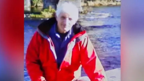 North Wales Police Brian Perry pictured wearing a bright red jacket and blue fleece underneath. He has grey hair and a river is visible behind him.