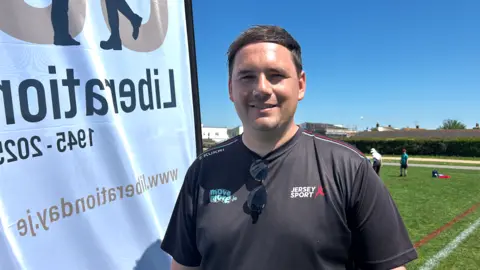 Man in black T-shirt. He has black hair and black sunglasses. A white flag is flying behind. Green pitch behind him.