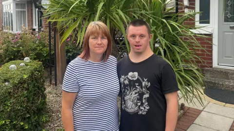 Alison Freeman A picture of Alsion Freeman (left) who has light brown shoulder length hair and is wearing a striped top and her son Lewis (right) who has shaved dark hair and is waring a black t-shirt with a design on the front. They are standing in front of a house with trees and shrubs behind them.