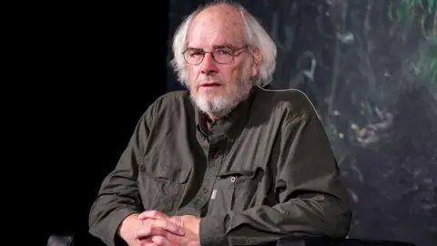 Jack Horner seated wearing a dark green shirt with hands crossed. He is wearing eye glasses and has white hair.
