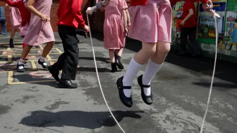 PA A girl wearing a red and white chequered school dress using a skipping rope jumps in the air. Children behind her are playing hopskotch, only their torsos can be seen. A mural showing a greengrocers can be seen behind them. 