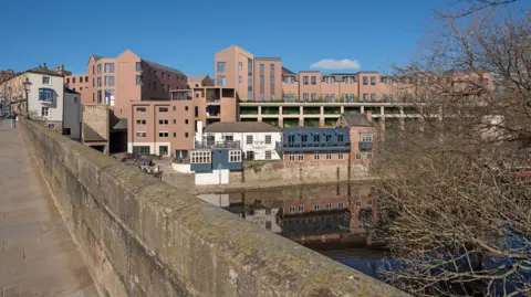 Computer generated image of how the redeveloped Prince Bishops site could look, featuring the River Wear in the foreground partially hidden behind a tree. The image shows the red brick impression of how the new blocks behind the existing riverside buildings. A stone bridge wall over the river acts a leading line towards the new development.