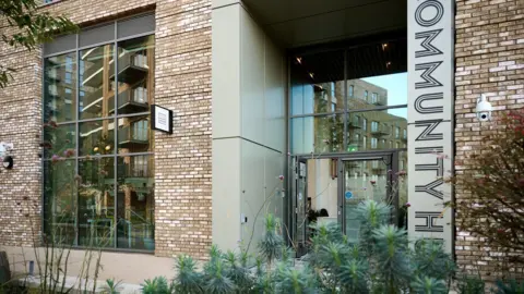 THP Photography An external view of the hub - a brick and plaster building with big glass doors and windows and a large black and beige vertical sign reading 'community hub'. Green plants sit in the foreground