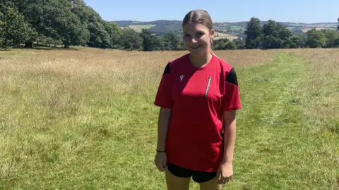 A girl with tied-back blond hair wearing a red running top stands in a green field 