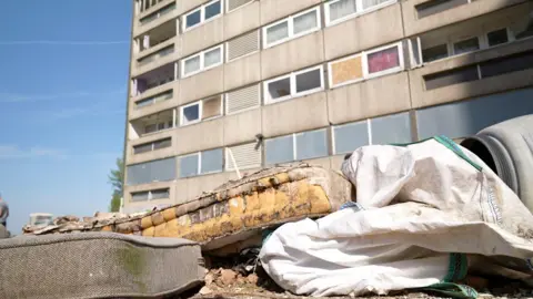 A dirty cushion, mattress and sheet lie on the ground in front of a block of flats.