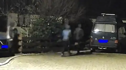 Devon and Cornwall Police CCTV image shows three individuals in hoodies walking near a parked vehicle. It is dark. The individuals are facing away from the CCTV.