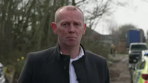 A man with short light brown hair and wearing a white shirt and a dark coat is being interviewed near to the site. The side of a police car and a lorry can be seen nearby.