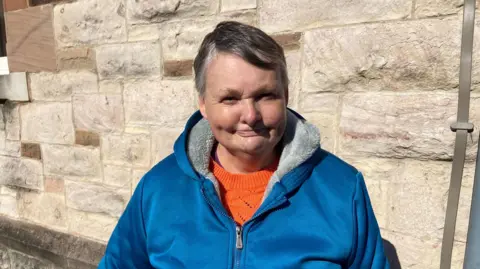 Janice Wylie in a blue hoodie on Larne Main Street with a cream wall in the background.