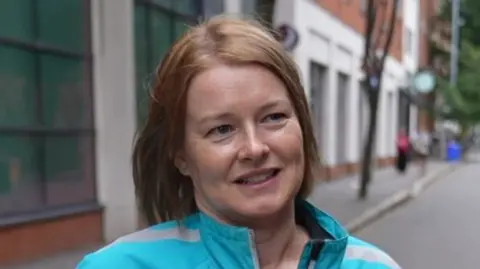 Anne has short ginger hair and is smiling at the camera as she looks to her left. She has a blue coat on, and behind her is brown and white buildings.