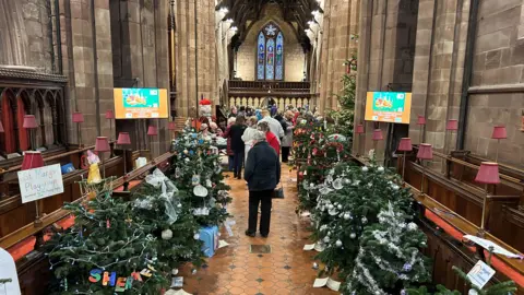 Several people walking down an avenue of decorated Christmas trees in a church with a number of pews on either side