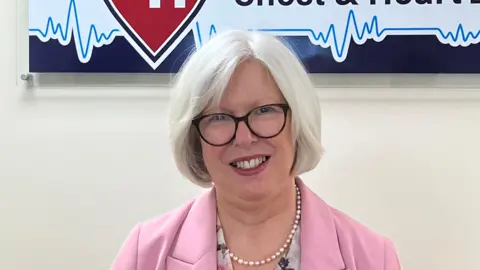 A grey haired woman with glasses and a pink suit smiles and looks down the camera
