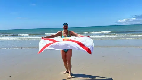 Jersey Long Distance Swimming Club Committee A woman holding a Jersey flag on the beach. Blue skies.