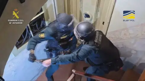 Two police officers wearing helmets and body armour using a battering ram to break through a door. They are standing in a narrow hallway. The back of their uniform has "GUARDIA CIVIL" written on it.