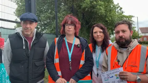 A man on the left is wearing a cap and a black top and the woman second from the left with glasses has red hair and is wearing blue and red. The next woman has brown hair and is wearing a white top, while the man on the right has dark hair and a beard. Three people, apart from the man on the left, are also wearing organge hi-vis jackets. 