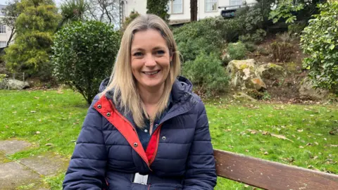 Ellie Lawrence, of Carers Guernsey, is wearing a navy jacket while sitting on a bench and smiling at the camera