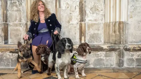 Pictoria A woman with blonde curly hair sits against a wall at Gloucester Cathedral as she holds onto four dogs.