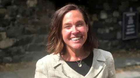 BBC A woman in a white blazer and black top smiling at the camera. She has brown hair.