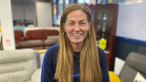 BBC Louise is smiling at the camera. She has long, straight ginger hair and is wearing a navy training top. She has gold hoop earrings.