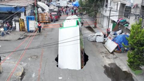 A large white truck is falling into a sinkhole, cordoned off by red tape, on a street in Mexico City.