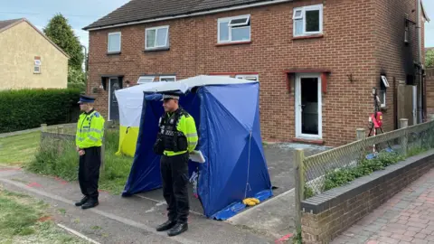 Two uniformed police officers stand guard, with a couple of police tents erected on the front drive of a semi-detached house. There is black charring to the side of the house.