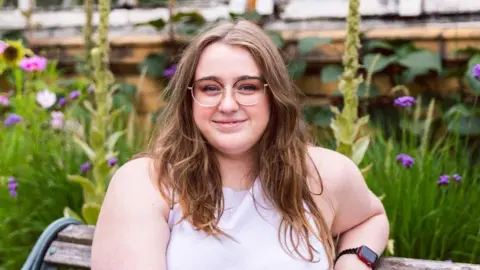 Eleanor Cotton Cotton sitting on a bench with greenery in the background. She wears an apple watch and purple dress. She has long brown hair and wears thin, clear-framed glasses