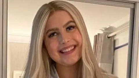 A woman with long blonde hair in a room with pale walls