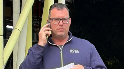 David Stroud, 44, after pleading guilty to harassing a woman on a train because of her sex. He is wearing a blue zip-up top, glasses, jeans and is holding a letter in one hand and a phone to his ear with the other. 