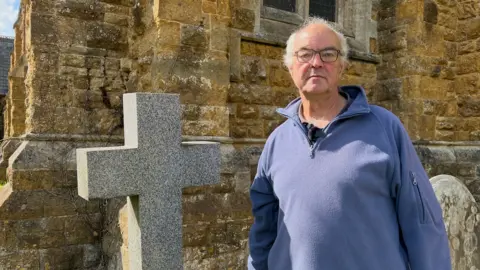 Philip Wallis, a man with short white hair, is standing next to a grey stone cross. Behind him are is a church, built of sand-coloured bricks. He is wearing glasses and a blue fleece.