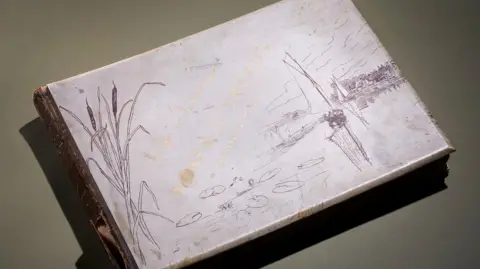 Roseberys View of the cover of 'Life and Landscape on the Norfolk Broads' by Peter Henry Emerson. The title is written in gold ink and the white paper has drawings of reeds and water symbolising the Norfolk Broads.