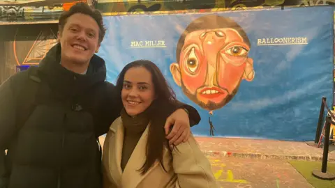 Conor Grovestock with his sister Olivia at the Mac Miller Balloonerism screening in London. Conor poses with his arm around his sister's shoulders in front of album artwork painted on a wall - the artwork depicts a cubist-style portrait of Mac's head as a balloon with a small figure holding it up. 