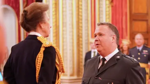 Princess Anne stands with her back to the camera as she talks to John Caldwell, who is wearing a police uniform.