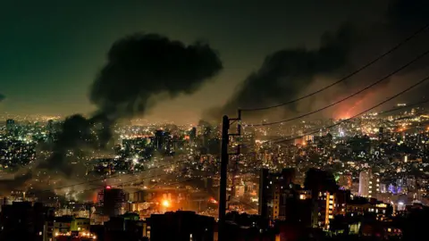 Tehran's skyline at night on Sunday 15 June, with three large plumes of smoke rising above the city and two large fires blazing below