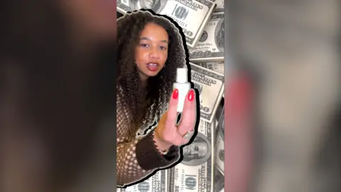 A woman with brown hair wearing a brown cardigan holds up a white bottle of correction fluid against a backdrop of American bank notes.