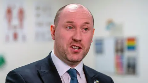 PA Media Health Secretary Neil Gray, wearing a dark suit, pink shirt and navy tie, is pictured in a medical setting - posters on the walls behind him blurred.