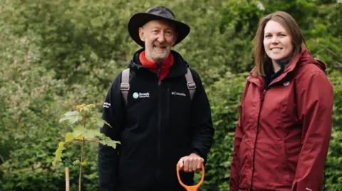 Broads Authority John Packman and Lynsey Skeates standing next to the sapling at the How Hill Nature Reserve on the Broads. The pair are both stood to the right of the plant. John Packman is wearing a black coat with a red scarf and has a hat on his head. He is looking directly at the camera and smiling and has a shovel with an orange handle in his hand. On his right is Lynsey Skeates who is wearing a red coat and she is looking directly at the camera and smiling.