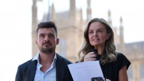 PA Media Laura Enston (centre), and Stephen Beard, the grandchildren of Ruth Ellis, making a statement to media outside the Houses of Parliament. Enston has shoulder length, dark blonde hair and wears a short-sleeved black dress with purple nail varnish. She is holding a piece of paper with her statement written on it.