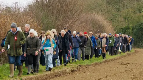 Block East Green Solar A group of people, some with dogs, walk along the edge of a field.