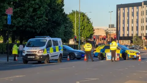 Lis Henderson / BBC View of the crash. A police car and van are parked across a road with a silver car in the middle. A number of police officers are stood around in yellow high visibility jackets.