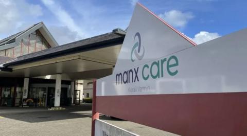 A large white sign with a pink base with Manx Care written on it outside the entrance to Noble's Hospital. The entrance is fronted by a canopy held up by while pillars. The letting on the sign in grey and green and has the Manx translation of Manx Care written beneath it.