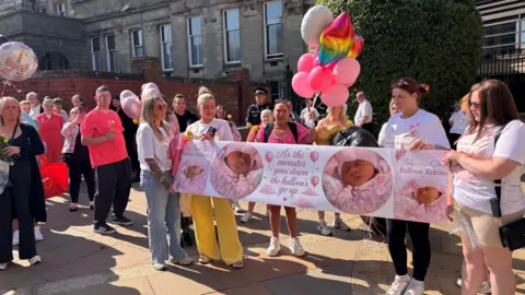 A group of people gathered outside a court, holding a banner and balloons. The banner reads As the monster goes down the balloons go up, with pictures of Thea Wilson on it