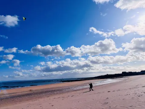Joanne MacLeod A wide sandy beach under a bright blue sky with scattered clouds. A person walks along the shore while a colourful kite flies high above.